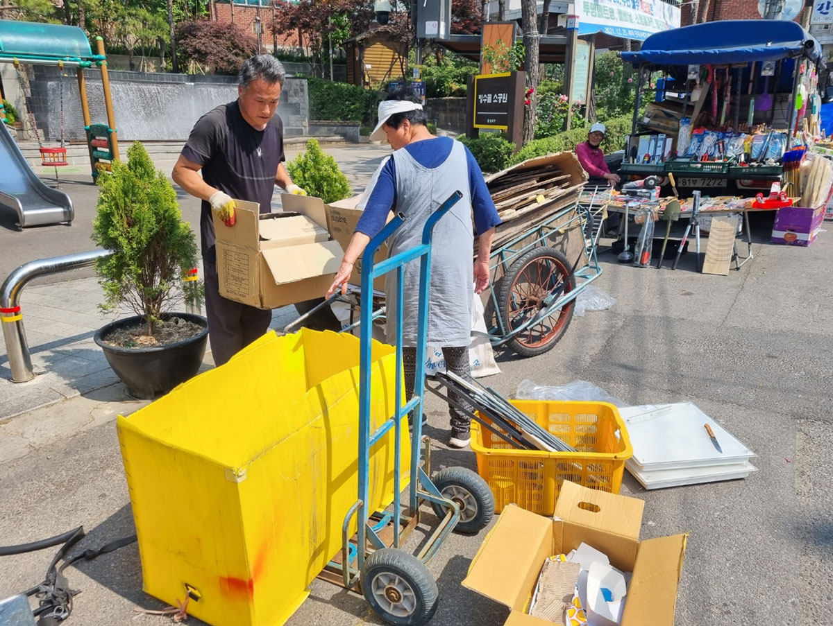 A man holding a cardboard box and a woman with her cart of recyclables.