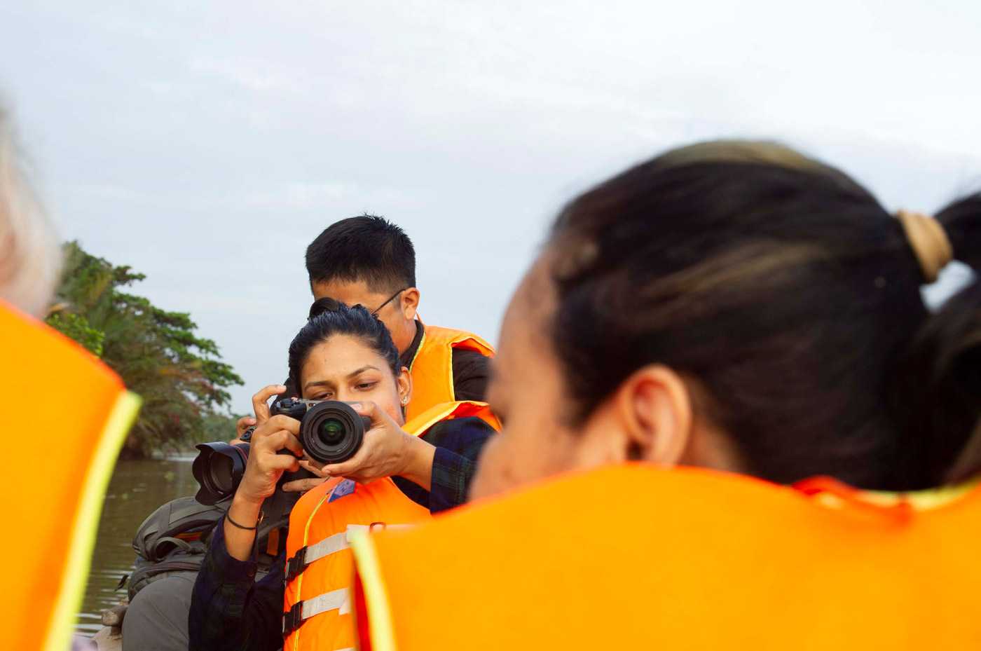 People in a boat wearing life jackets, one is aiming a camera forward.