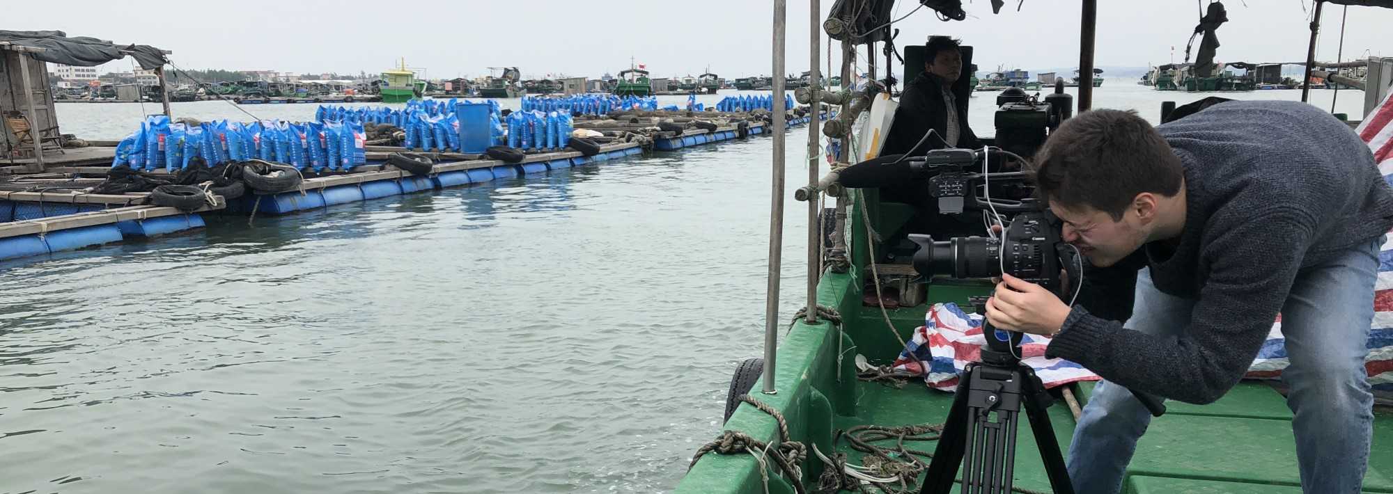 A man with a camera, crouched over on a boat near a fish farm in China.